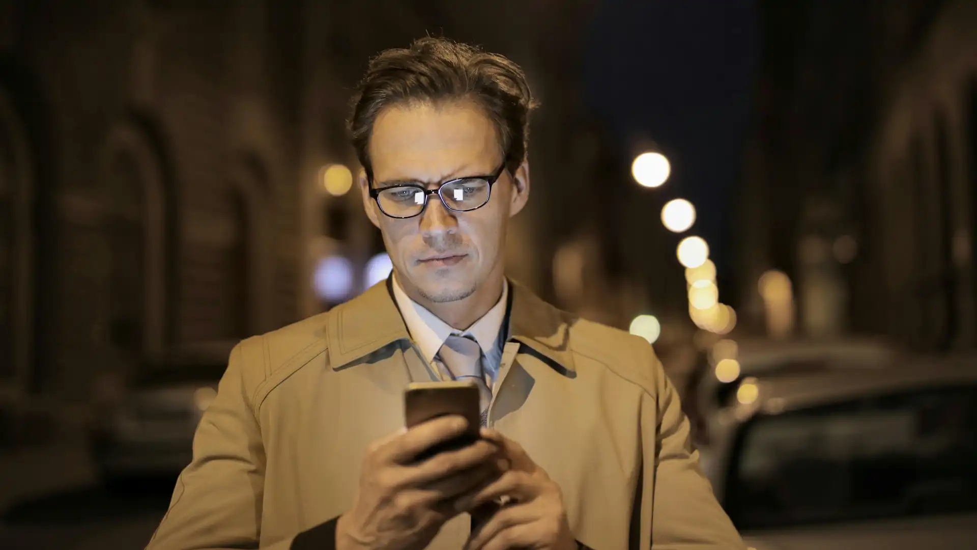 Man in a beige trench coat and glasses looking at his phone at night.